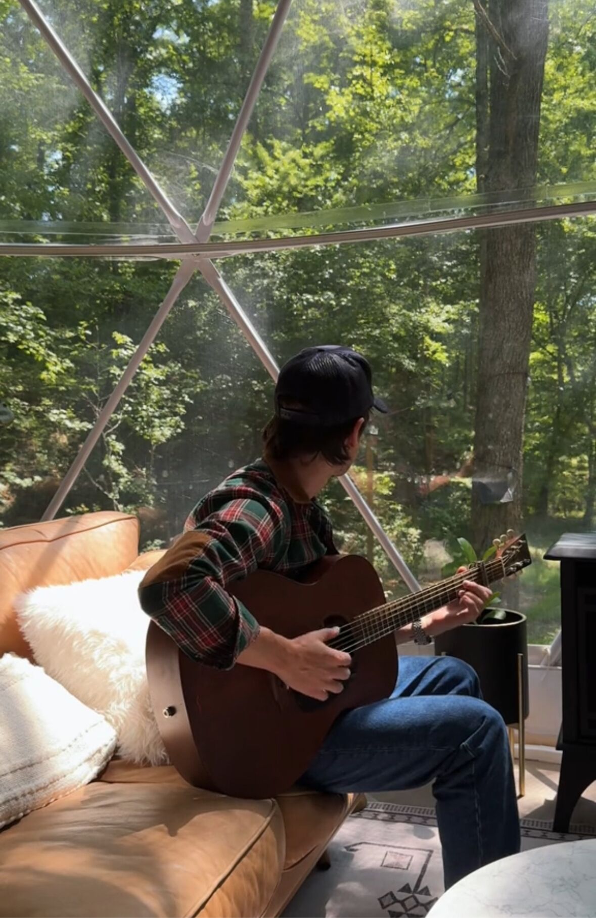 Una persona asseguda en un sofà de cuir tocant la guitarra en una habitació amb un gran finestral que donava a un bosc.