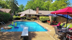 Une piscine dotée d’un espace de détente comprenant des chaises et des parasols, entourée de verdure et bordée d’un bâtiment avec une cheminée.