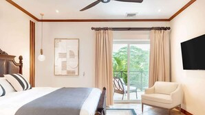 A bedroom with a bed, a ceiling fan, a mounted TV, a balcony with a chair, and a view of greenery outside. - Stylish Apartment w/ Hotel Amenities @ Los Sueños (Playa Herradura)