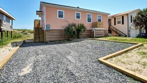 A two-story building with a wooden deck, a gravel driveway, and a small garden area. - West End Gulls~ Guest Favorite ~ Beach Access ~ Astonishing Views (Oak Island)