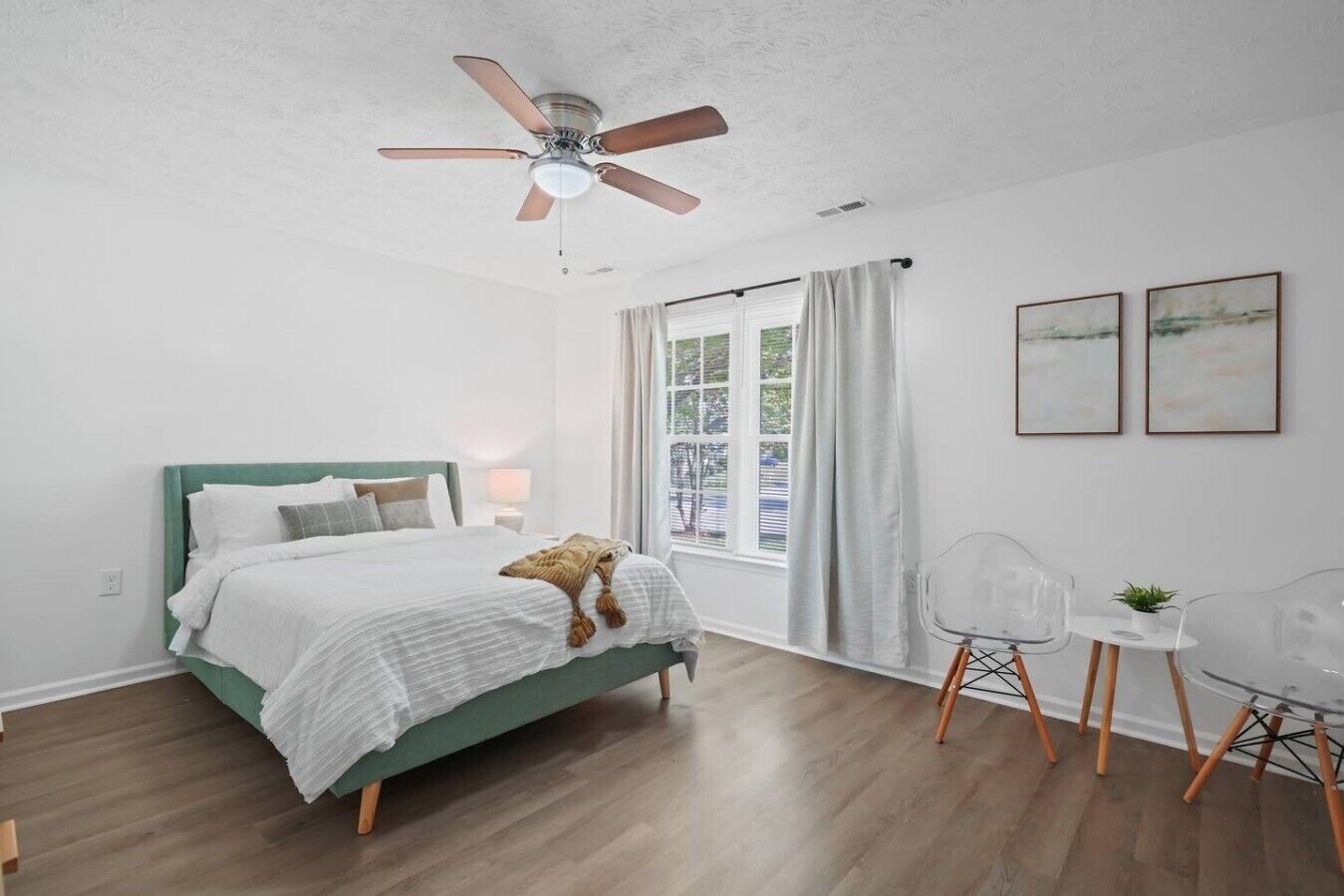 A bedroom with a bed, a ceiling fan, a window with curtains, and a small table with chairs.