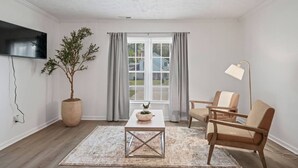 Una sala de estar con televisor, planta en maceta, mesa, dos sillas, lámpara de pie y ventana con cortinas.