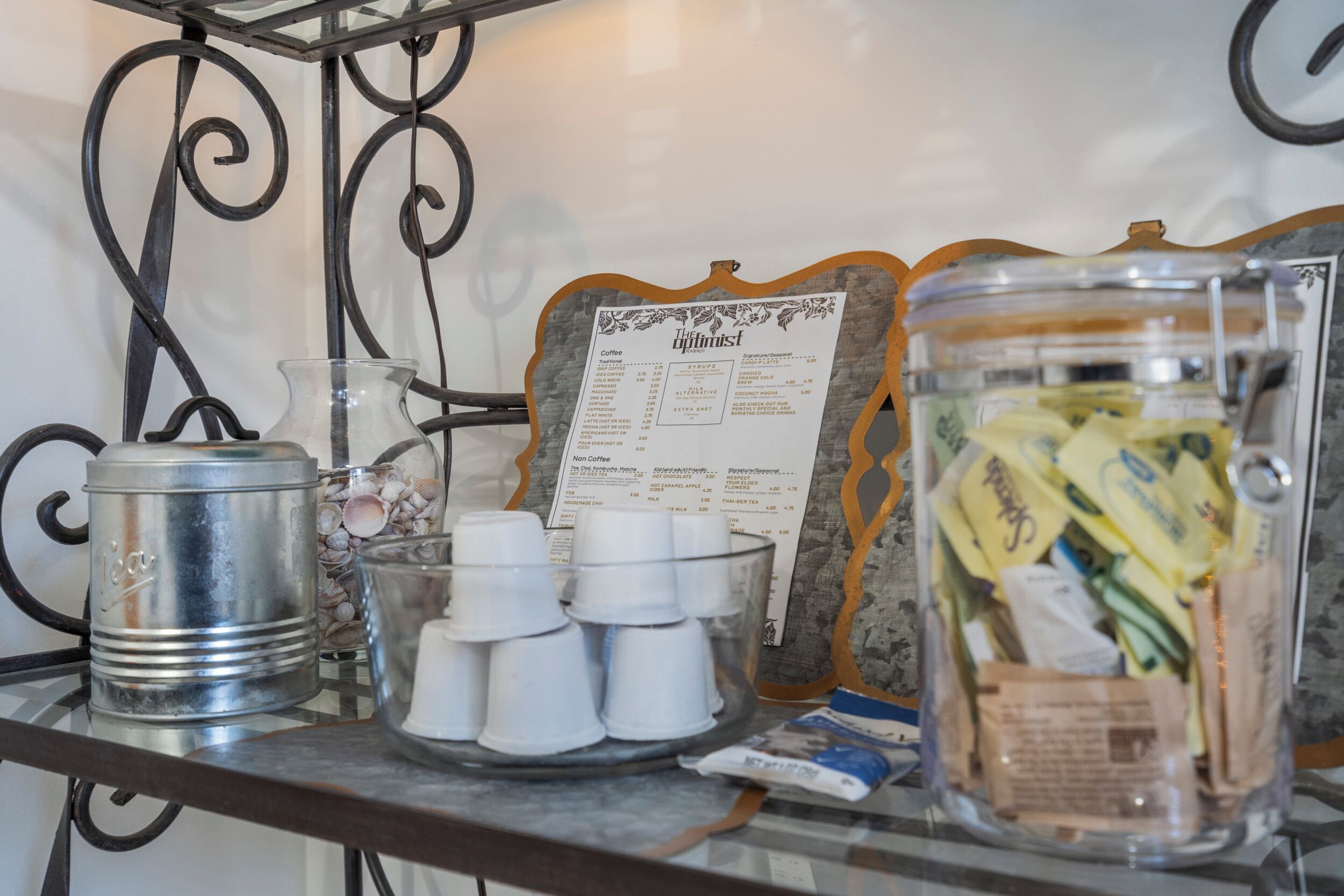 A coffee station with a menu, a metal container, a glass pitcher, disposable cups, and a jar of snacks.