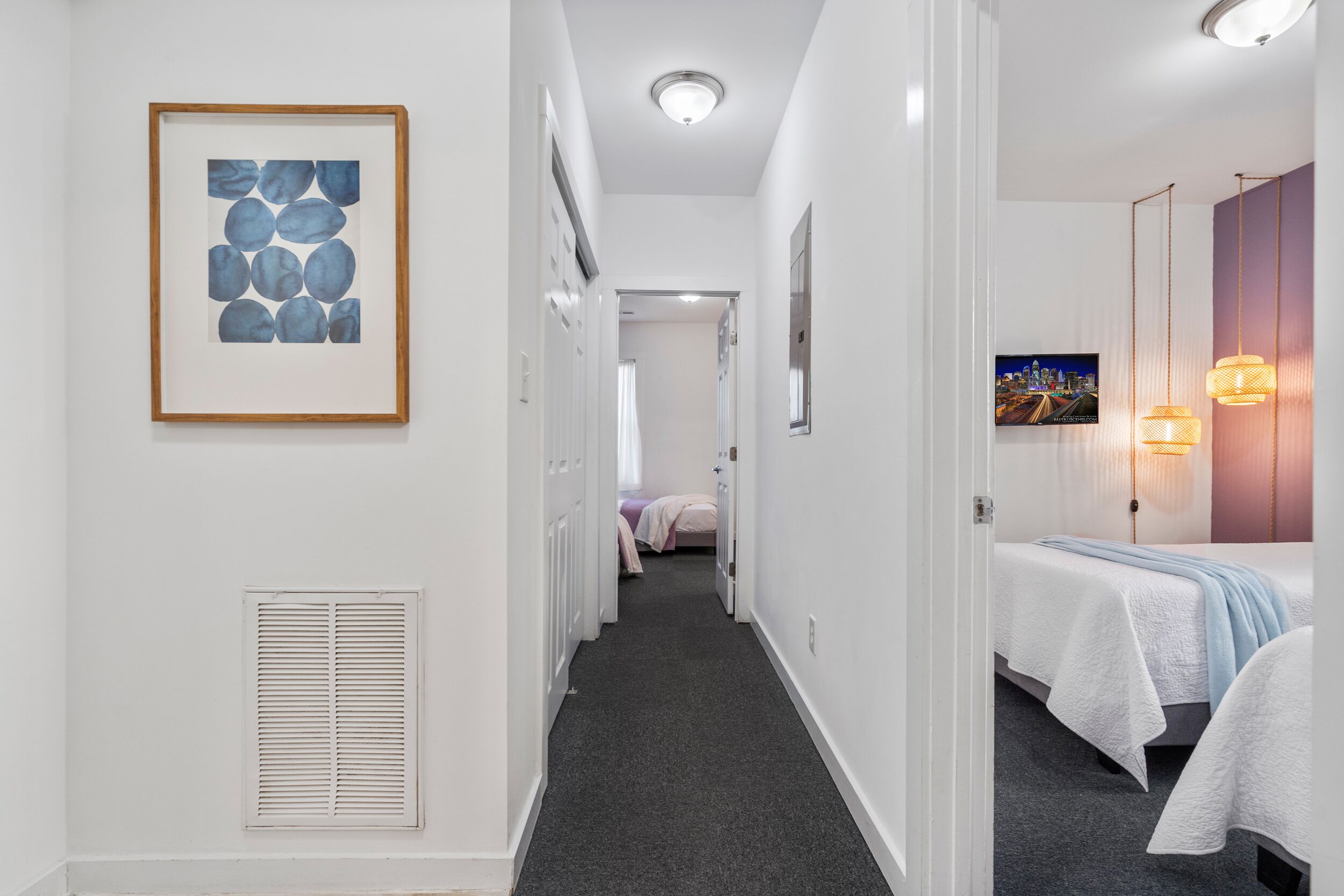 Pasillo de hotel con una obra de arte enmarcada, una cama con colcha blanca, un televisor en la pared y un pasillo que conduce a otra habitación.