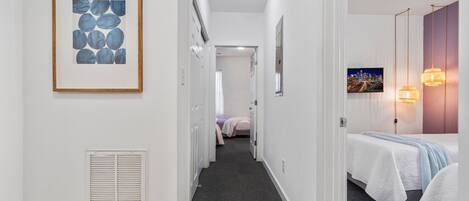 Pasillo de hotel con una obra de arte enmarcada, una cama con colcha blanca, un televisor en la pared y un pasillo que conduce a otra habitación.