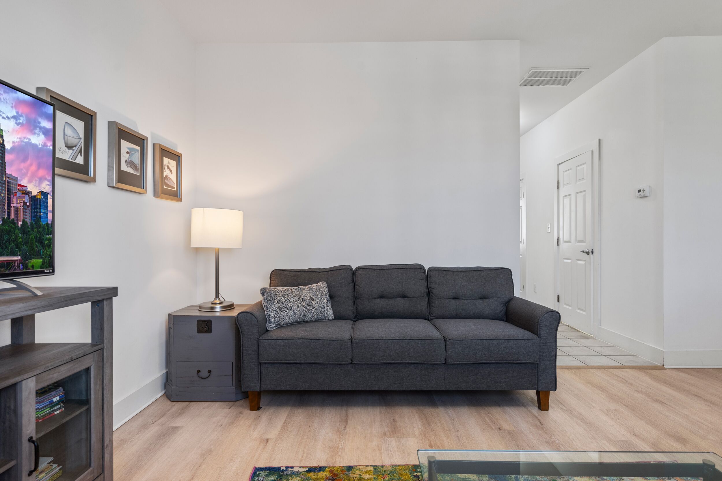Un salon avec un canapé gris, une table d’appoint avec une lampe, un téléviseur posé sur un meuble en bois et des tableaux encadrés accrochés au mur.