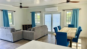 A spacious living room with a large wooden dining table, blue upholstered chairs, and a grey sectional sofa. There are ceiling fans, large windows with blue curtains, and an air conditioning unit on the wall. - Sands of Time is ready for You  (Rainbow Bay)