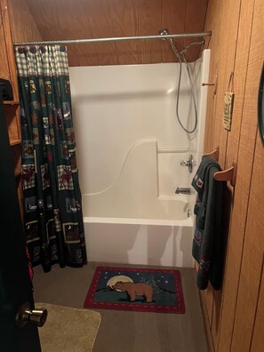 Un baño pequeño con paneles de madera, una bañera blanca, una ducha y una cortina con un diseño de oso. Se observa una puerta de madera con una toalla colgada.