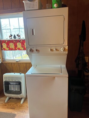 Cocina compacta con refrigerador blanco, estufa blanca y un pequeño calentador. Se ve una ventana con vista a árboles y una cortina roja decorativa.
