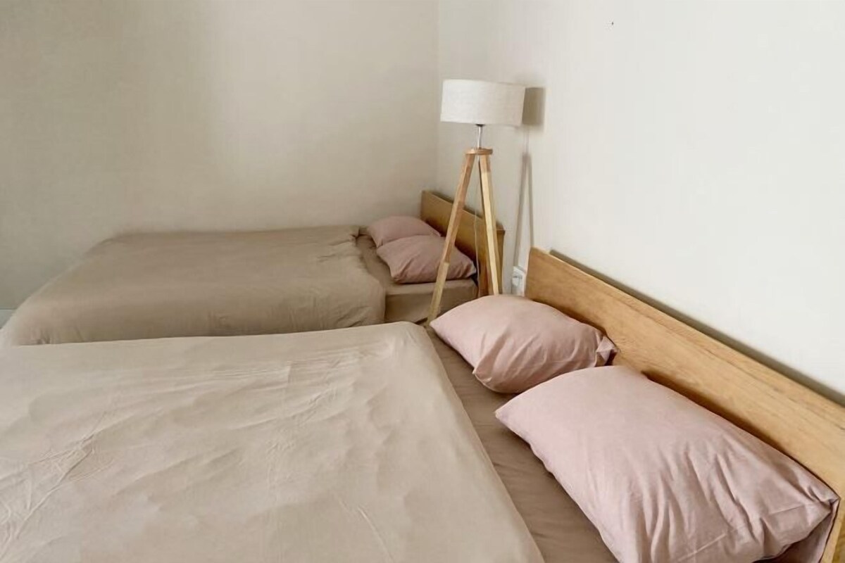 Una habitación de hotel con dos camas, cada una con una colcha color beige y una almohada rosa. Hay una lámpara de pie con pantalla blanca junto a una de las camas.
