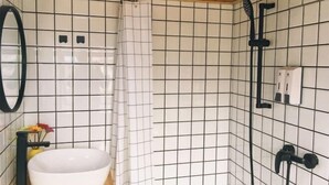 Ein Badezimmer mit weiß gefliesten Wänden, einem weißen Waschbecken, einem schwarzen Wasserhahn, einem Spiegel und einem Duschbereich.