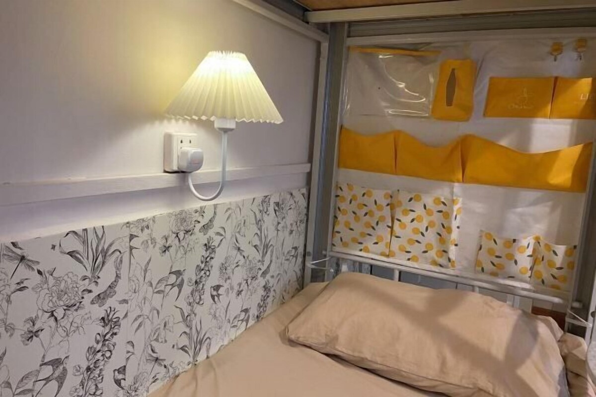 A hotel room with a floral wallpaper, a bedside lamp, a window with yellow curtains, and a bed with a beige cover.