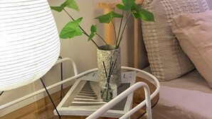 Ein weißer Stuhl mit einer Glasvase, in der eine Pflanze und Schilfrohre stecken, eine weiße Lampe und ein Bett mit einem gemusterten Kissen.
