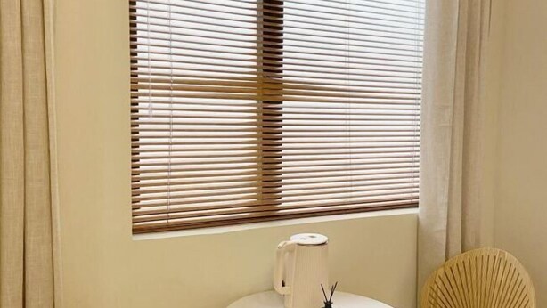 Ein Zimmer mit einem runden Tisch, einem Stuhl, einem Fenster mit Jalousien und einem kleinen weißen Gegenstand auf dem Tisch.