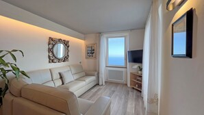 A living room with a beige sofa, a large mirror, a framed picture, a TV, and a window with a view of the sea. - Casa Camelia: your luxury apartment in the heart of Cinque Terre (Volastra)