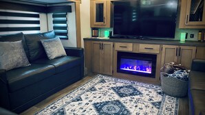 A modern living room with a black leather sofa, a flat-screen TV, wooden cabinets, a fireplace, and a patterned rug. - Secluded riverfront RV 7 Miles from Glacier National Park! (Coram)