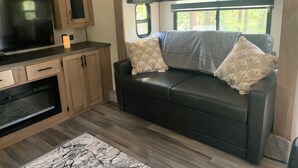 A compact RV interior with a kitchenette, a sofa with patterned cushions, and a large window providing a view of greenery. - Secluded riverfront RV 7 Miles from Glacier National Park! (Coram)