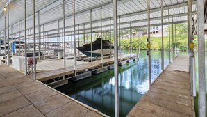 Eine überdachte Marina mit vertäuten Booten, einem Betonboden und einer Aussicht auf Grünflächen und Gebäude im Hintergrund.