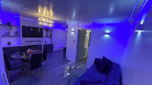 Una sala de estar moderna con una mesa de centro de vidrio, un sofá y un comedor con televisor y plantas.