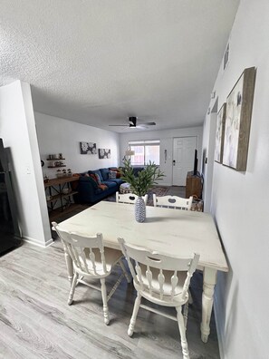 Un coin repas avec une table et des chaises blanches, un ventilateur de plafond et une vue sur le salon.