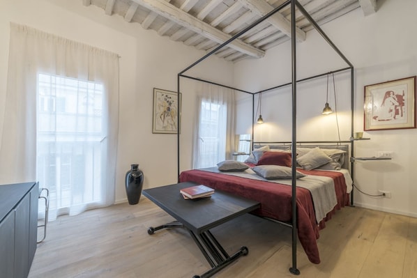 A bedroom with a four-poster bed, a bedside table with a book, a large vase, and a window with sheer curtains.