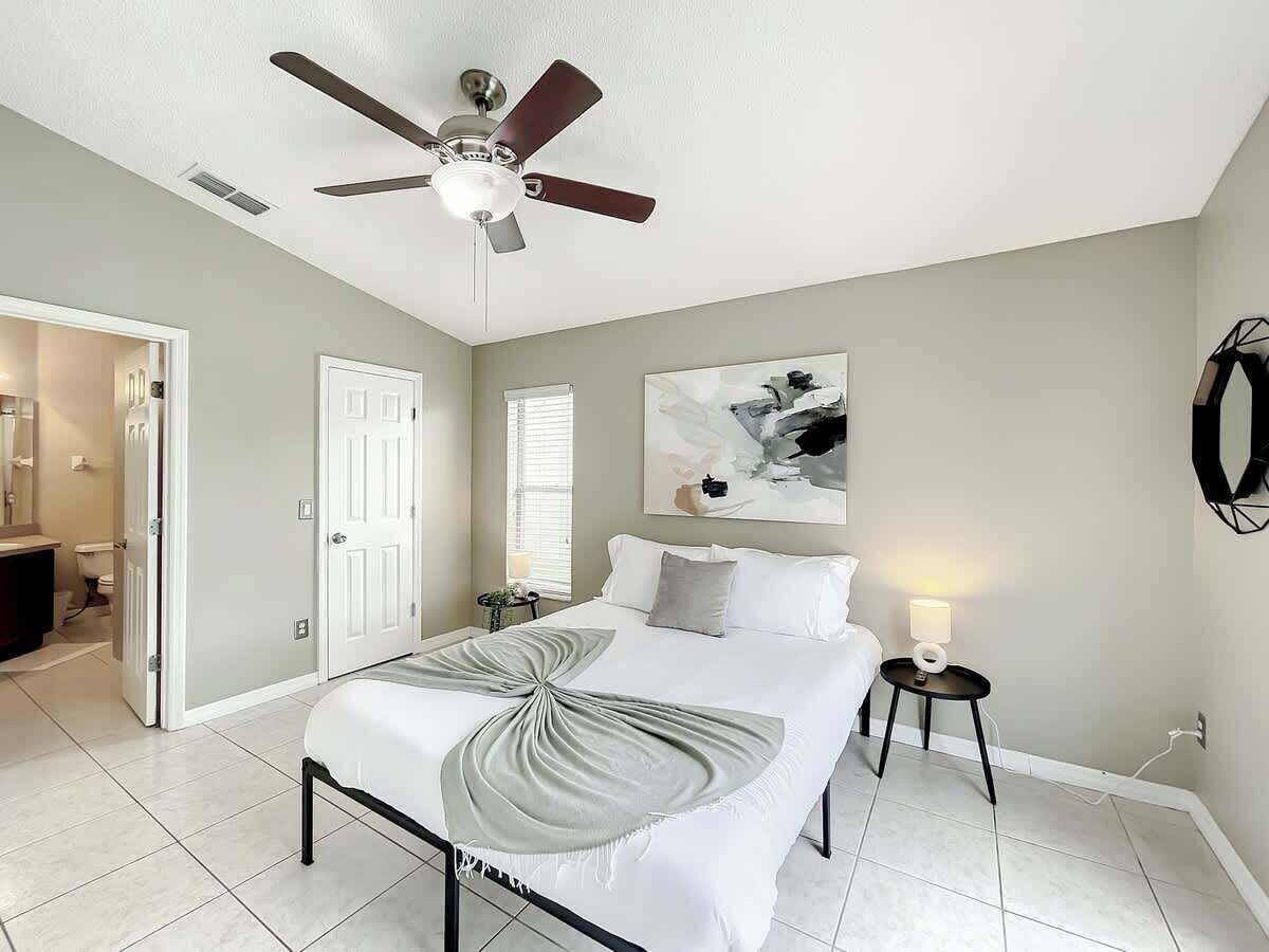 A bedroom with a bed, a ceiling fan, a bedside table with a lamp, and a painting on the wall.