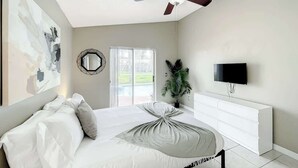 Une chambre à coucher comprenant un lit, un ventilateur de plafond, une télévision, une commode et une fenêtre donnant sur un paysage verdoyant.