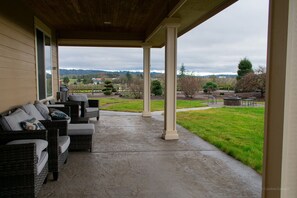 Un patio techado con mobiliario de mimbre y vistas a un jardín ajardinado y colinas al lejano.
