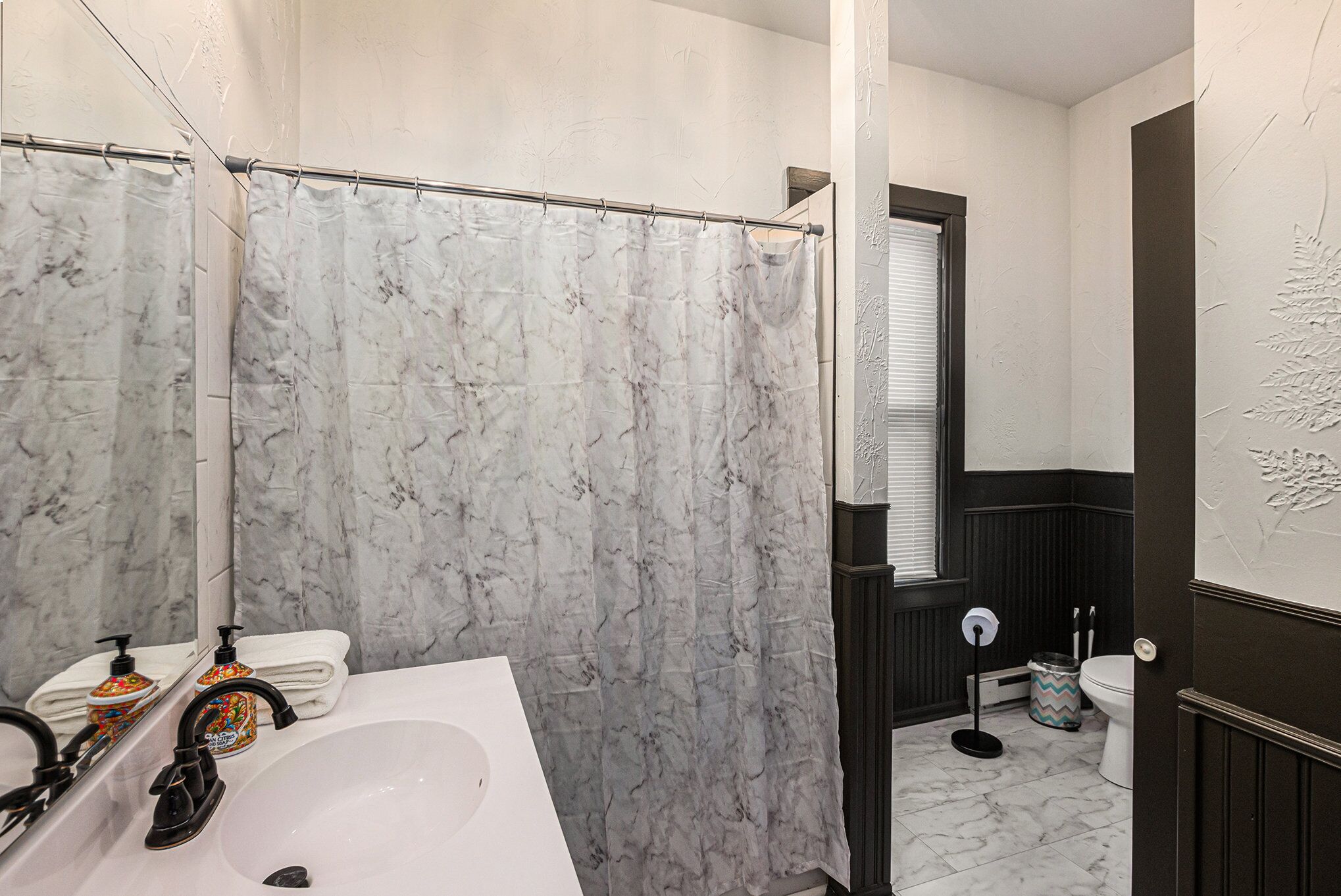 A bathroom with a white sink, a mirror, a shower curtain, a toilet, and a trash can.