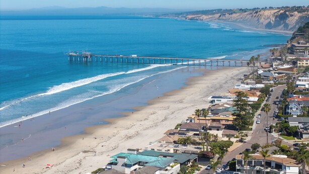 Un pueblo costero con un muelle que se adentra en el océano, propiedades frente a la playa y una vista despejada de la costa.