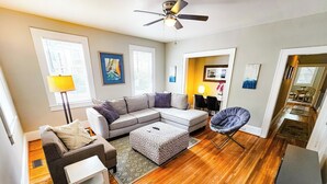 A living room with a sectional sofa, a small table, a chair, and a ceiling fan. - Modern Apt 86" TV! (Charlotte)