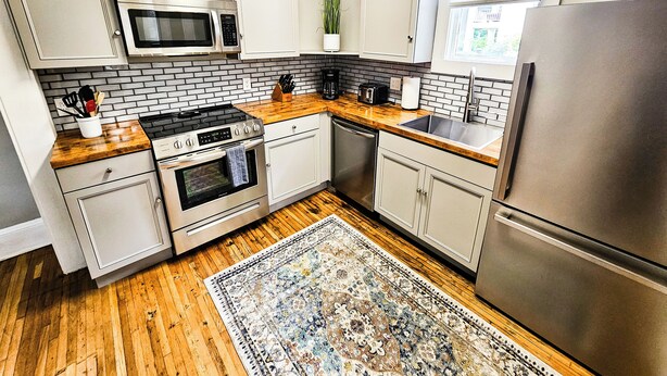 A modern kitchen with white cabinetry, a stainless steel refrigerator, a microwave, a stove, and a sink. - Modern Apt 86" TV! (Charlotte)
