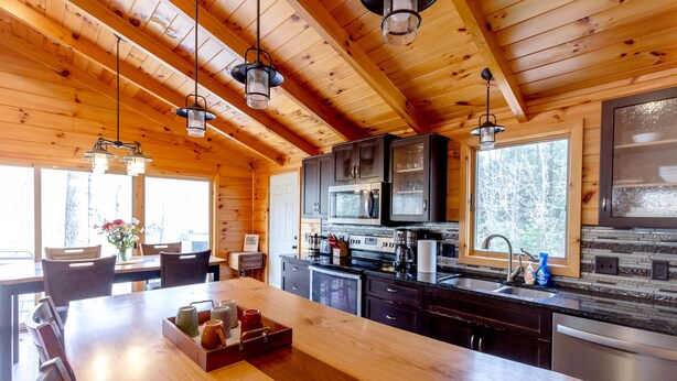 A wooden cabin interior with a large wooden dining table, kitchen area with appliances, and large windows. - Riverfront | Hot Tub, Fireplace, Kayaks & Tubes (Evart)