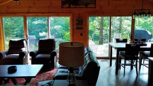 A cozy cabin interior with a sofa, chairs, a dining table, and a lamp. Large windows offer a view of the surrounding forest. - Riverfront | Hot Tub, Fireplace, Kayaks & Tubes (Evart)