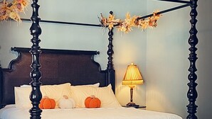 A four-poster bed with a dark wooden frame and a white bedspread. There is a lamp on a bedside table and a ceiling fan above the bed.