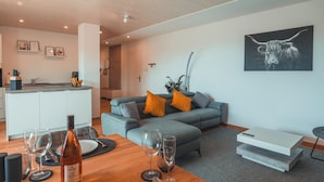 Een moderne woonkamer met een grijze bank, oranje kussens, een houten salontafel en een eethoek gedekt met borden, glazen en bestek.