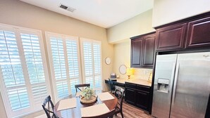 A dining area with a round table, chairs, and a potted plant. There is a kitchen with dark cabinets and a stainless steel refrigerator.