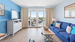 A modern living room with a blue sofa, a wooden coffee table, a white TV stand, and a balcony with outdoor furniture.