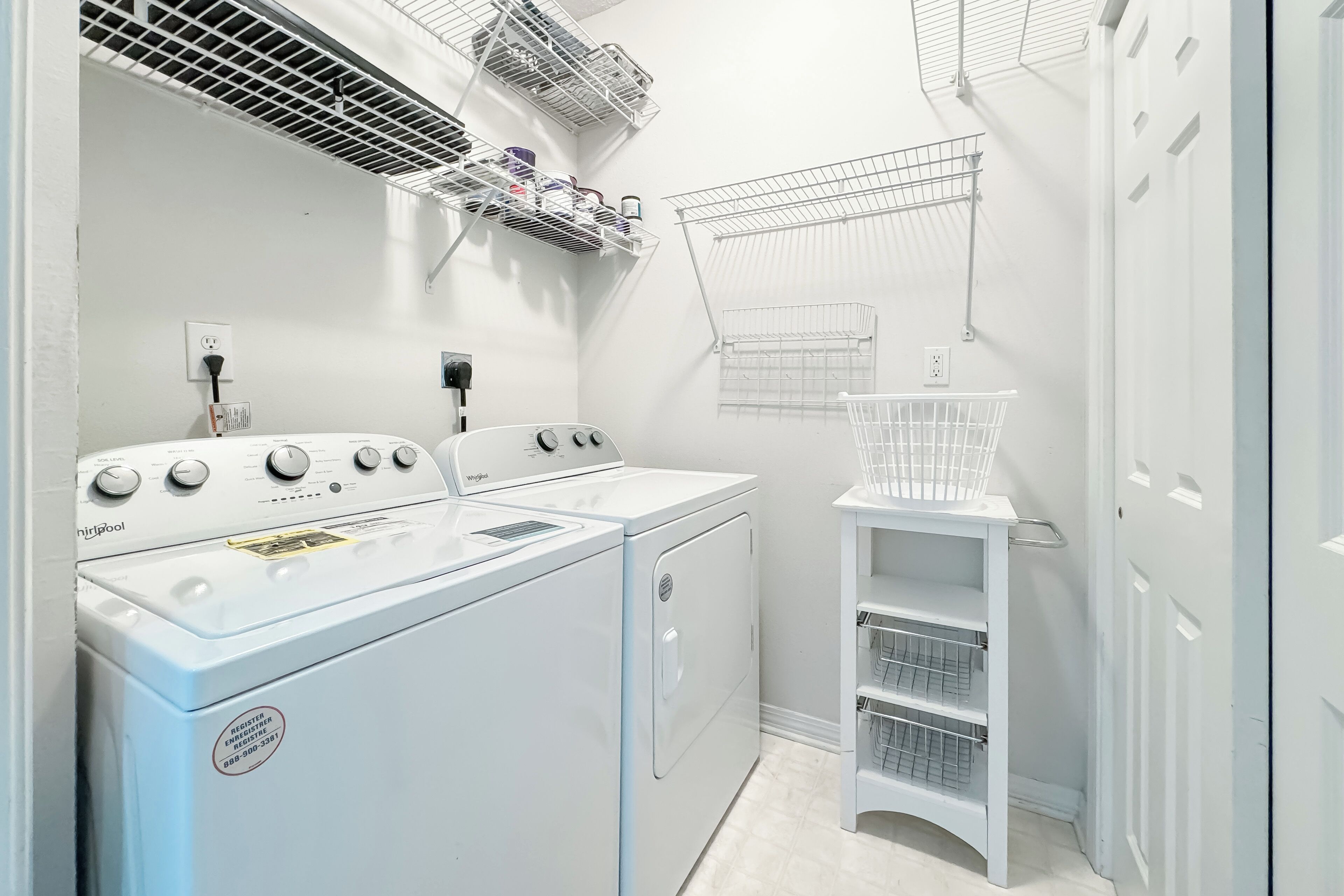 A laundry area with a Whirlpool washer and dryer, a white shelf with hanging clothes, and a white laundry basket.