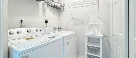 A laundry area with a Whirlpool washer and dryer, a white shelf with hanging clothes, and a white laundry basket.