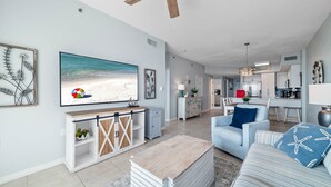 Een ruime woonkamer met een grote flatscreen-tv, een plafondventilator en een keuken op de achtergrond.