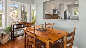 Un coin repas avec une table et des chaises en bois, une plante en pot et une vue sur la cuisine par une porte ouverte.