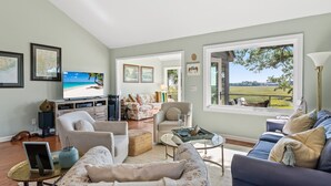 Un vaste salon doté d’une grande fenêtre donnant sur une plage et des champs, un coin salon confortable avec des canapés et des fauteuils, et un téléviseur à écran plat posé sur un meuble.