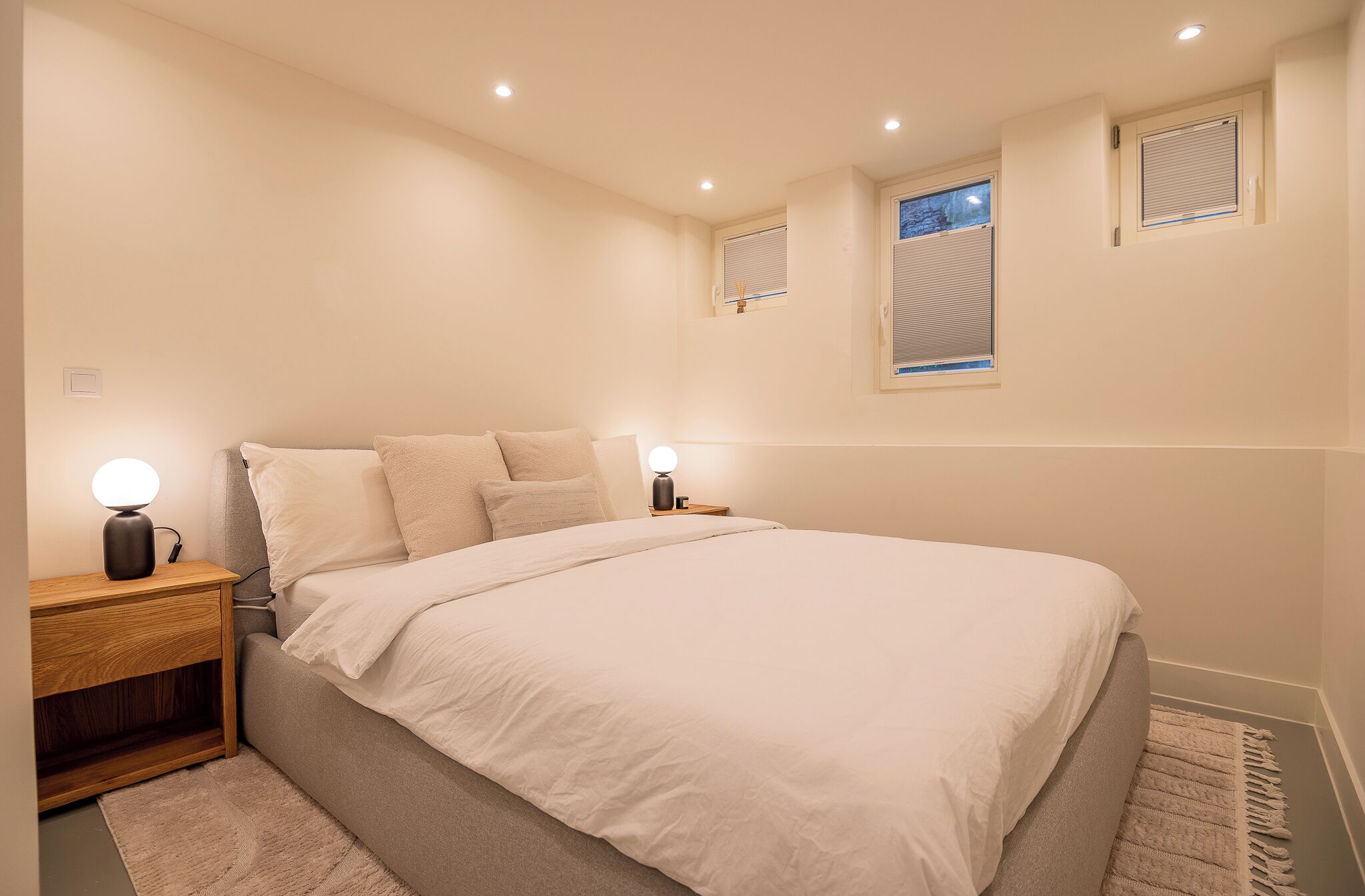 Une chambre à coucher avec un grand lit, deux tables de chevet avec des lampes, une fenêtre avec des stores et une petite étagère au-dessus de la fenêtre.