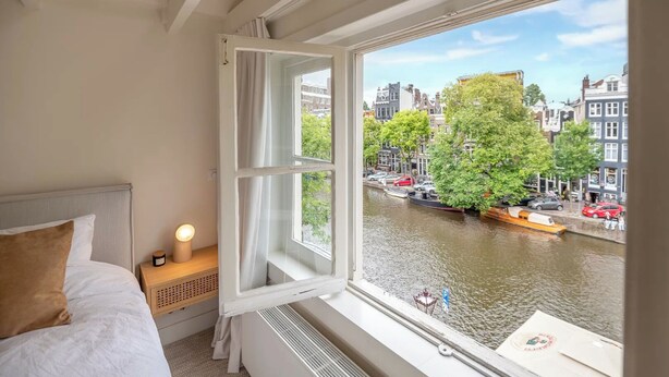 A hotel room with a bed, a bedside table with a lamp, and a window overlooking a canal with boats and buildings. - Elegant, sophisticated and unique canal house part of the famous canal district (Amsterdam)
