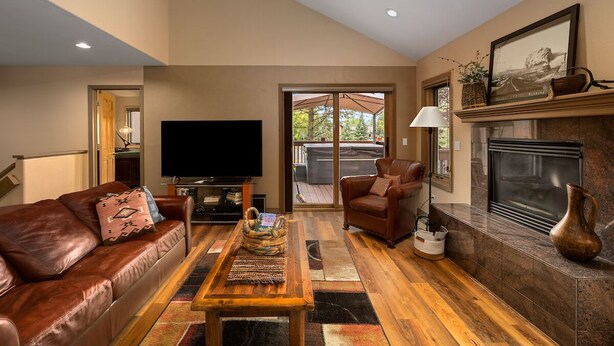 A spacious living room with a leather sofa, a wooden coffee table, a flat-screen TV, a fireplace, and a large sliding glass door leading to an outdoor area. - A Ducking Good Time! AC, Large Deck with Hot Tub & 2 Car Garage! (Steamboat Springs)