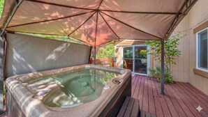 A hot tub under a canopy, wooden deck, and a sliding glass door leading to a patio. - A Ducking Good Time! AC, Large Deck with Hot Tub & 2 Car Garage! (Steamboat Springs)