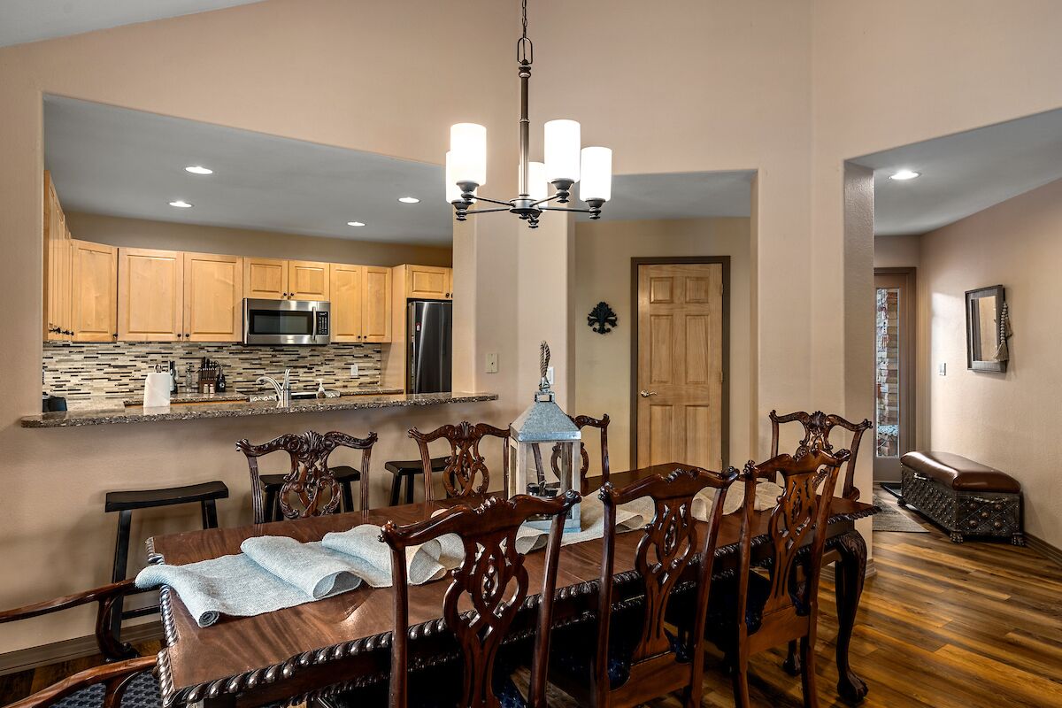 A dining area with a wooden table and chairs, a chandelier, and a kitchen in the background.