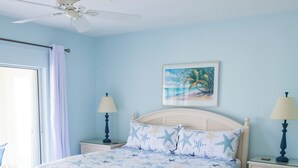 Une chambre à coucher comprenant un lit, deux tables de chevet, un ventilateur de plafond, un tableau et une vue sur l’extérieur.