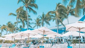 Un complexe hôtelier en bord de mer, avec des rangées de chaises longues blanches et de parasols bleus, des palmiers et un ciel d’un bleu limpide.